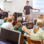 students sitting in classroom and teacher is standing while poiting towards screen