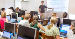 students sitting in classroom and teacher is standing while poiting towards screen