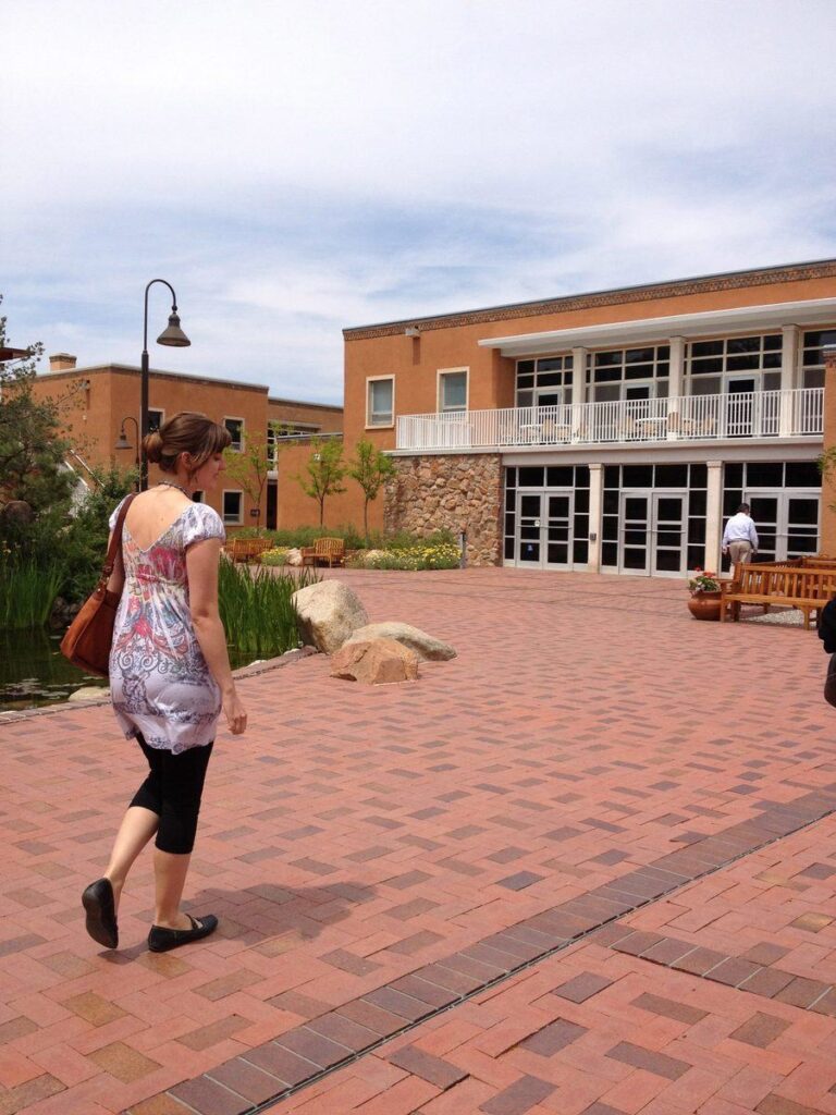 A woman with a brown bag walks on a brick path toward a modern, tan building with large windows and white balcony railings under a partly cloudy sky. Another person stands near a wooden bench.