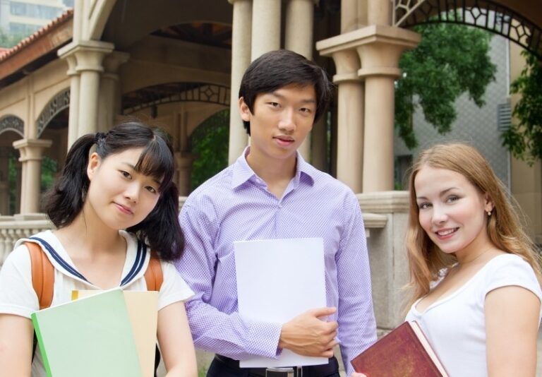 Three students stand outdoors in front of a building, holding books and notebooks. They are facing the camera and smiling, appearing relaxed and friendly. The setting includes columns and greenery in the background.