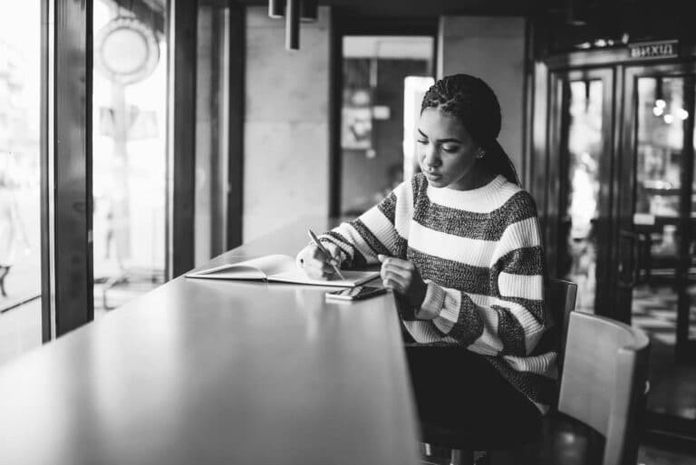 A woman sits at a counter by a window, writing in a notebook with a pen. She is wearing a striped sweater and appears focused. The setting is a modern, well-lit indoor space, possibly a café. The image is in black and white.