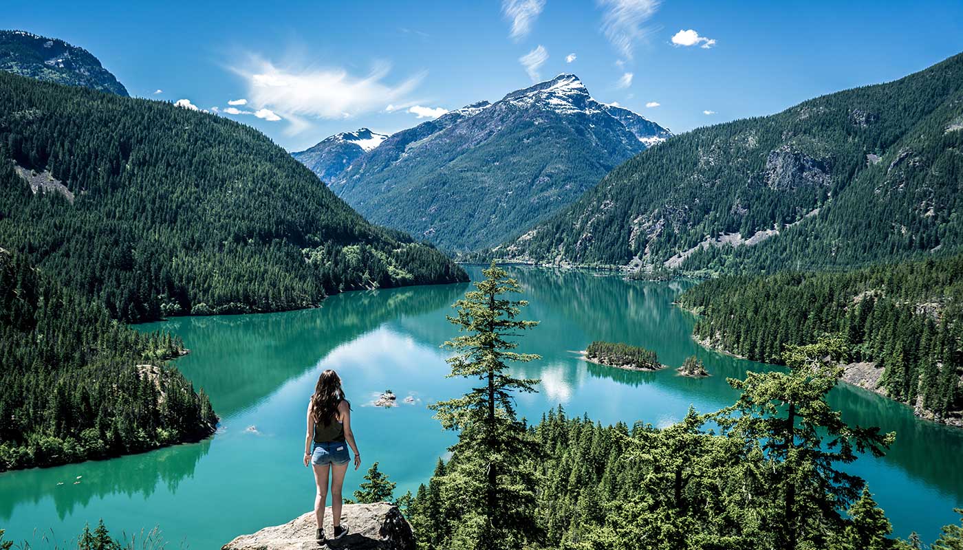 A person stands on a rocky ledge overlooking a turquoise lake surrounded by dense evergreen forests and snow-capped mountains under a bright blue sky.