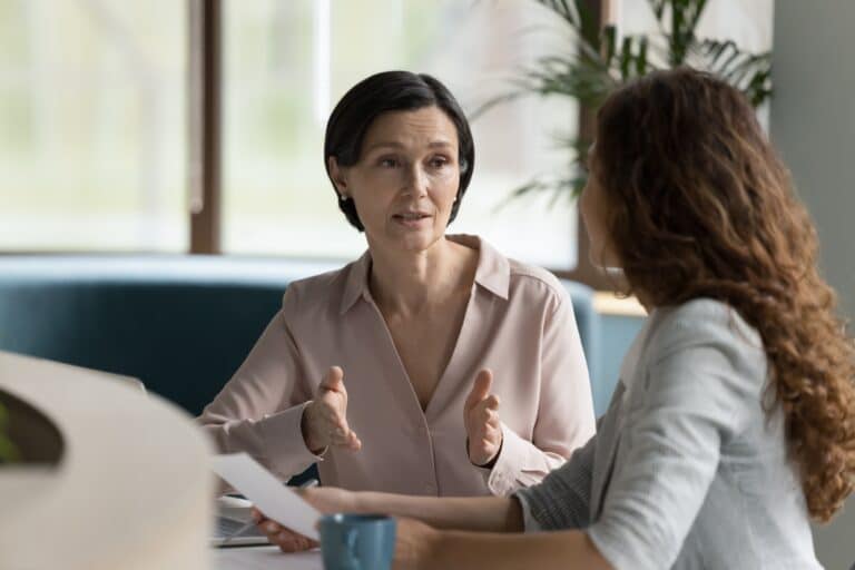 Two women sit at a table having a serious conversation; one gestures with her hands while speaking, and the other listens, holding a blue mug and a piece of paper. A window and plants are in the background.