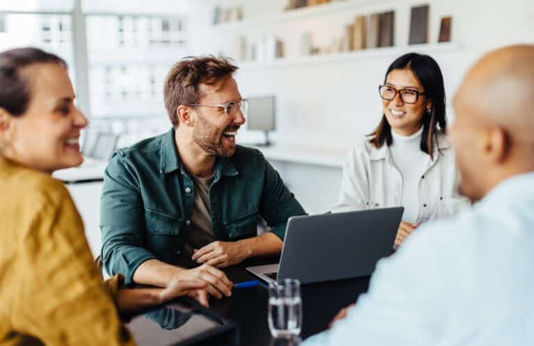 Four people sit around a table in a bright office, smiling and talking. A laptop and a glass of water are on the table, and books line shelves in the background, suggesting a casual meeting or discussion.