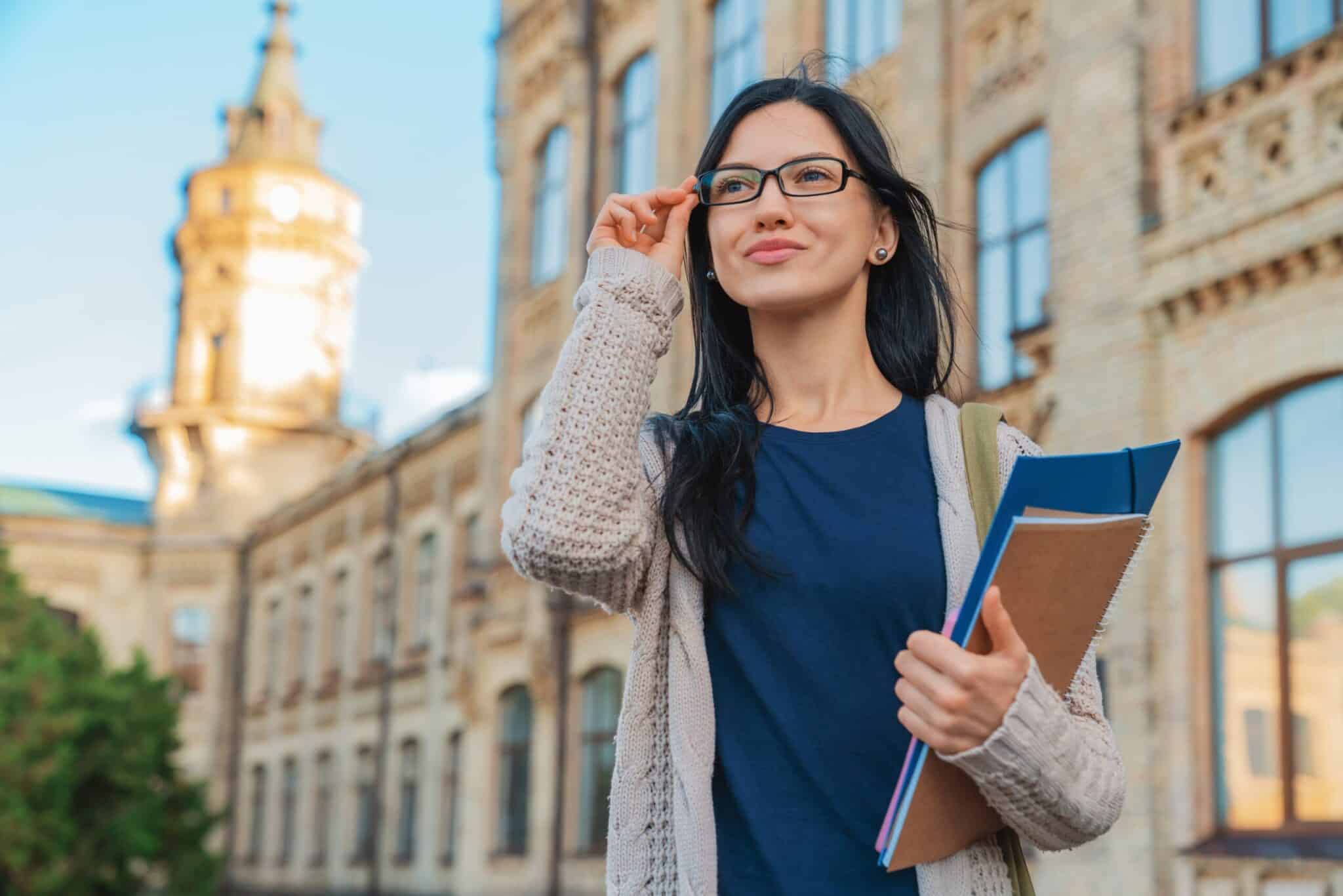 A young woman with long dark hair and glasses stands outside a historic building, holding notebooks and smiling, wearing a blue shirt and light sweater.