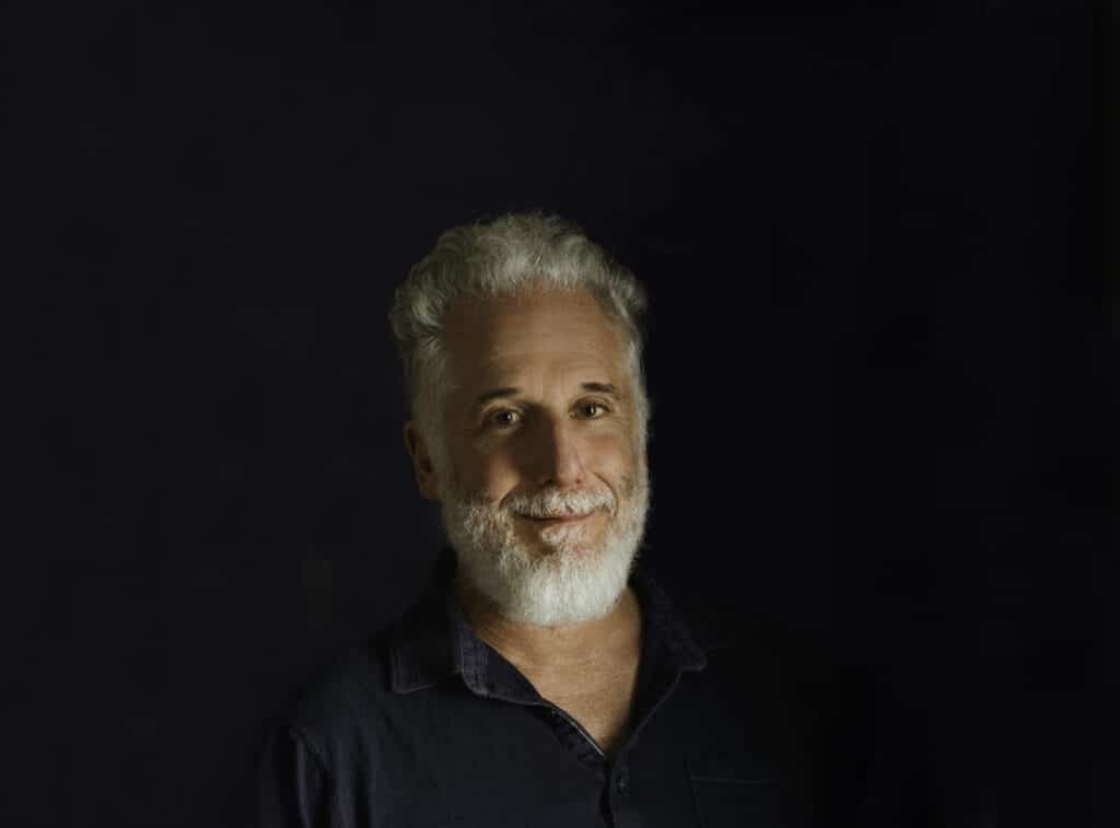 Headshot of a smiling man with gray hair and beard, wearing a dark shirt against a black background.