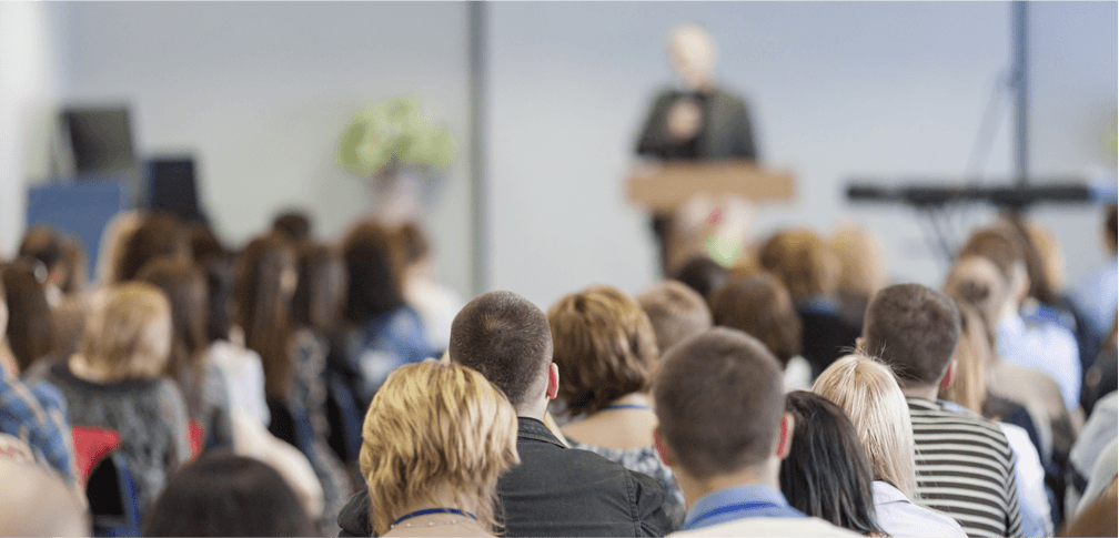 Audience listening to a speaker at a conference or seminar. Backs of heads visible, speaker blurred in background.