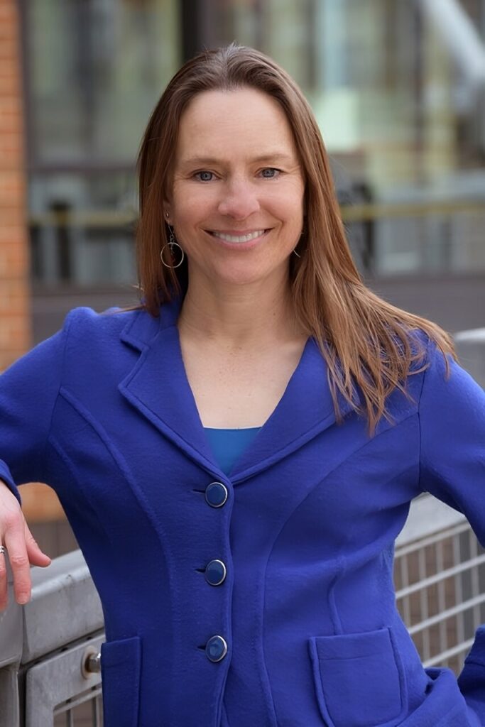 Headshot of a woman with shoulder-length brown hair wearing a royal blue blazer. She is smiling and leaning on a metal railing.