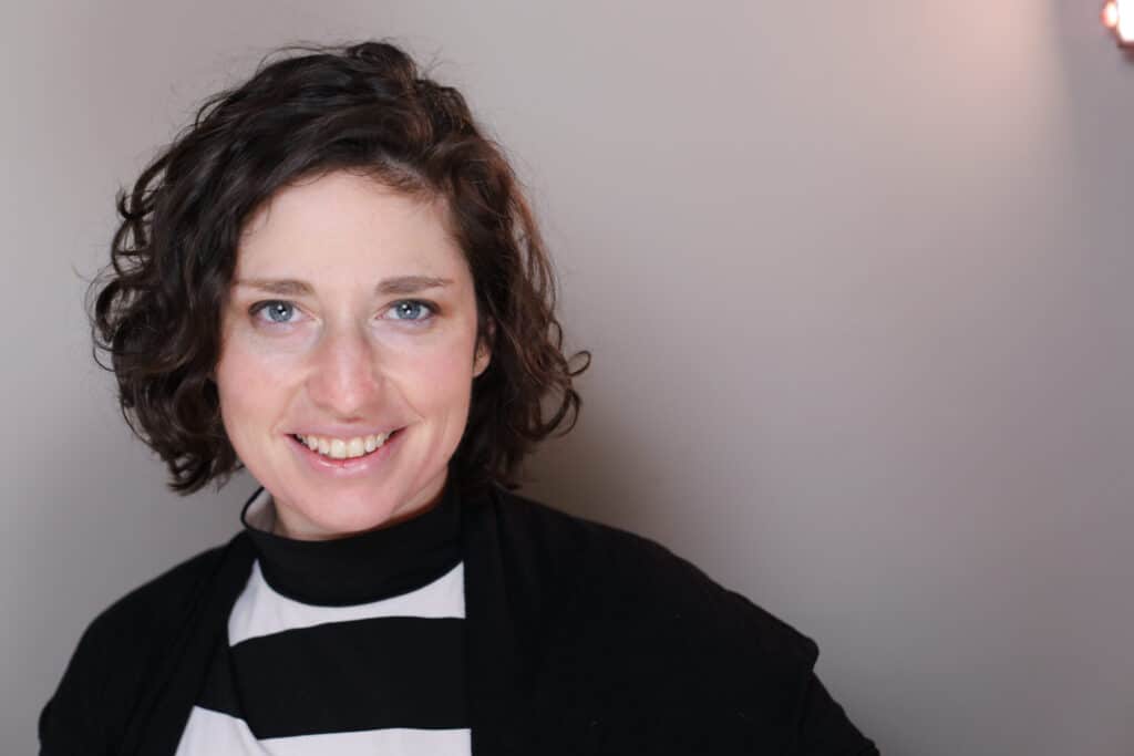 Smiling woman with curly brown hair wearing a black and white striped shirt and black cardigan against a light gray background.
