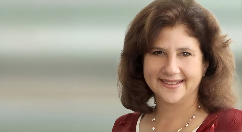 A woman with shoulder-length brown hair is smiling at the camera. She is wearing a red top and a necklace. The background is softly blurred with shades of green and cream.