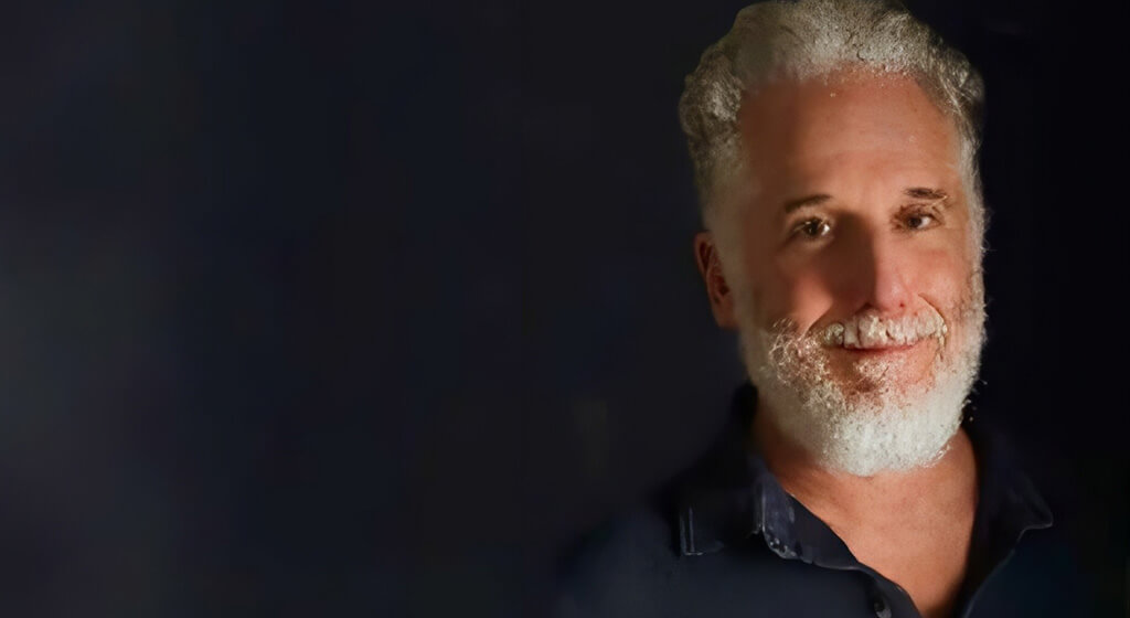 A man with curly gray hair and a beard smiles slightly while standing against a dark, plain background. He is wearing a dark shirt and is positioned on the right side of the image.