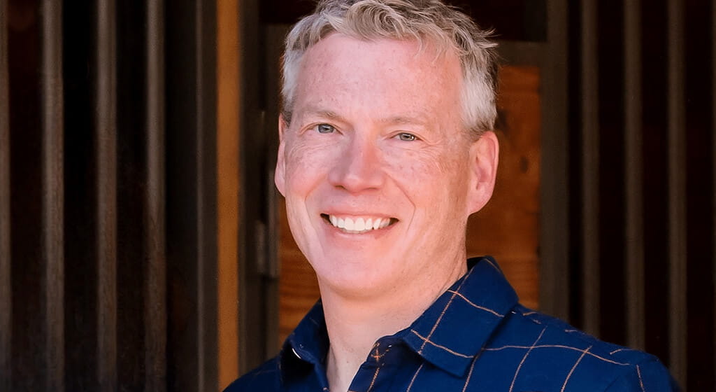 A man with short gray hair smiles at the camera, wearing a blue shirt with an orange grid pattern, standing in front of a wooden background.