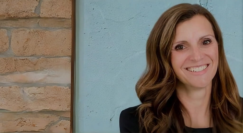 A headshot of a smiling woman with long, wavy brunette hair wearing a black top. She is positioned on the right side of the frame against a light blue textured wall, which meets a vertical section of light-colored brick on the left.