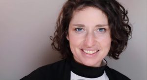 A woman with short, curly dark hair smiles at the camera. She is wearing a black and white top and standing in front of a plain light gray background.