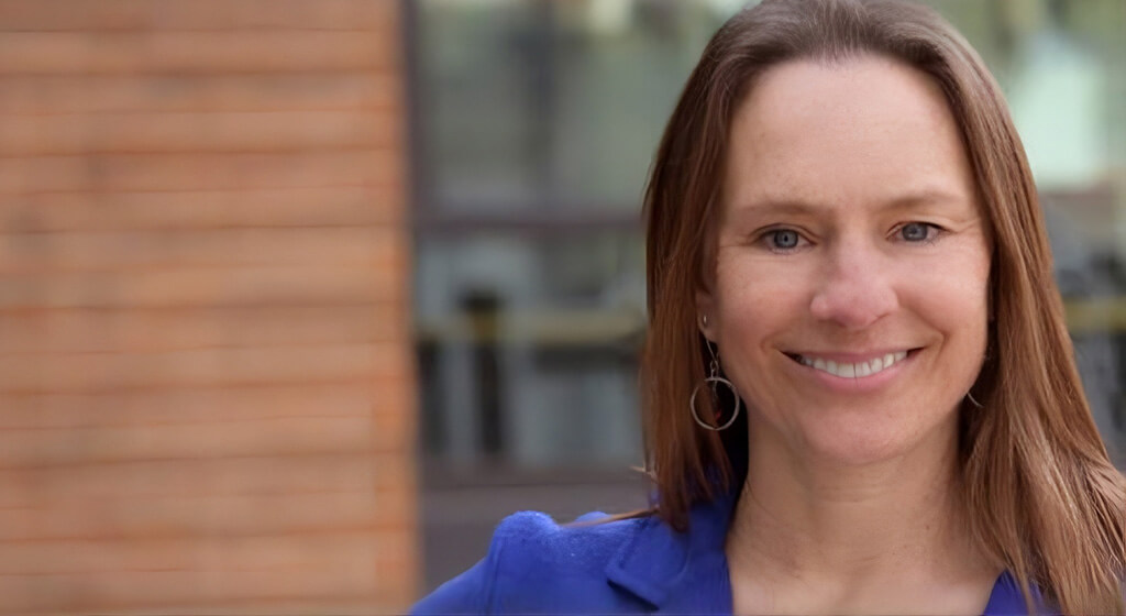 A woman with straight brown hair, wearing a blue blazer and hoop earrings, smiles at the camera. The background is blurred, showing a brick wall and a window.