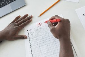 A person using a red pen to circle an answer on a standardized test sheet while resting their hand on the table. A laptop and a yellow pencil are visible in the background.