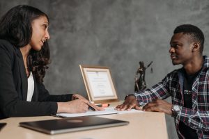 A professional woman and a man engaged in a serious discussion at a desk, with documents and a framed certificate visible. A small statue of Lady Justice stands nearby, indicating a legal context.