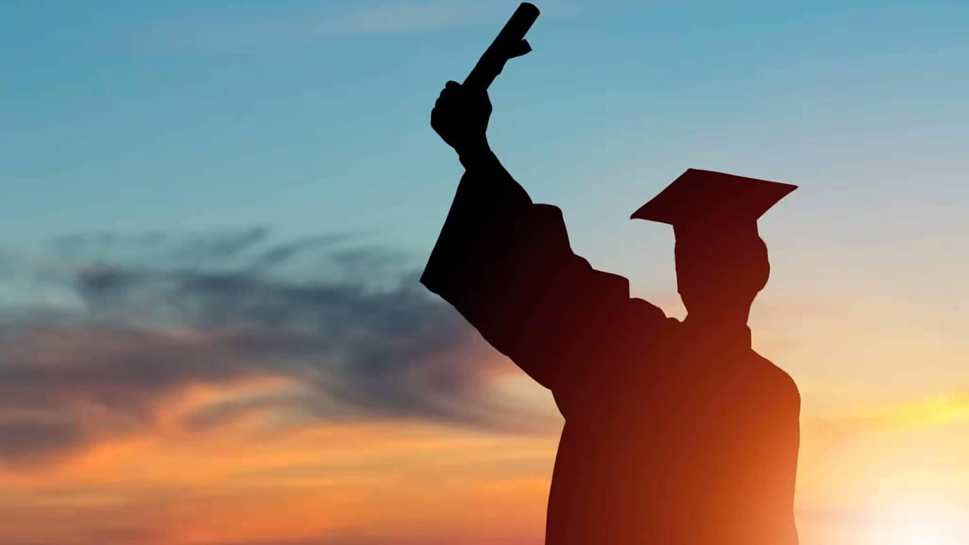 Silhouette of a graduate wearing a cap and gown holding up a diploma against a colorful sunset sky.