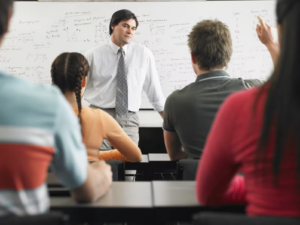 A confident instructor in a white shirt and patterned tie stands in front of a whiteboard filled with equations, engaging a classroom of attentive students. The students, wearing colorful attire, sit at sleek black desks, some raising their hands to participate, highlighting an interactive learning environment.