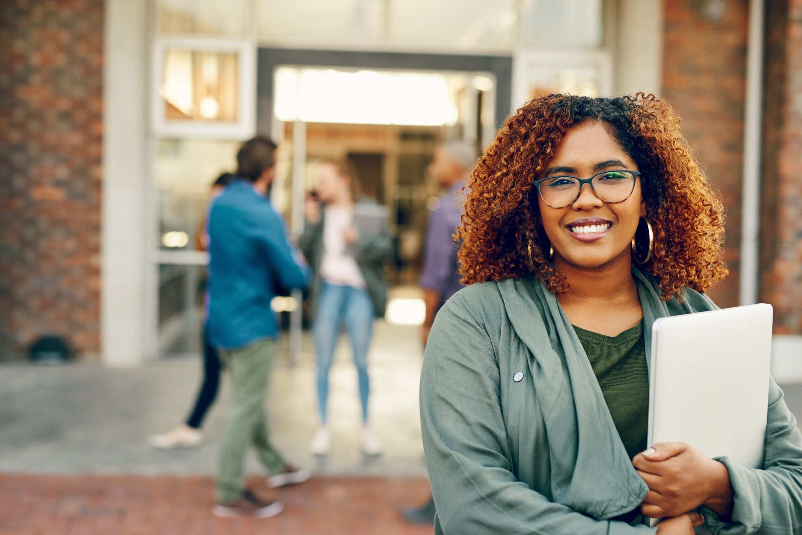 A woman with curly, shoulder-length hair and glasses stands confidently in the foreground, holding a laptop close to her chest. She wears a green jacket over a dark top, and the sun-kissed brick backdrop frames a lively group of people engaging in conversation.