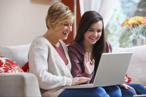 A cozy living room scene featuring two women sitting on a plush, light gray couch adorned with vibrant red-patterned cushions. The older woman, dressed in a soft cream cardigan, and the younger woman, in a burgundy hoodie, share a moment of joy as they engage with a silver laptop, sunlight illuminating the inviting space.