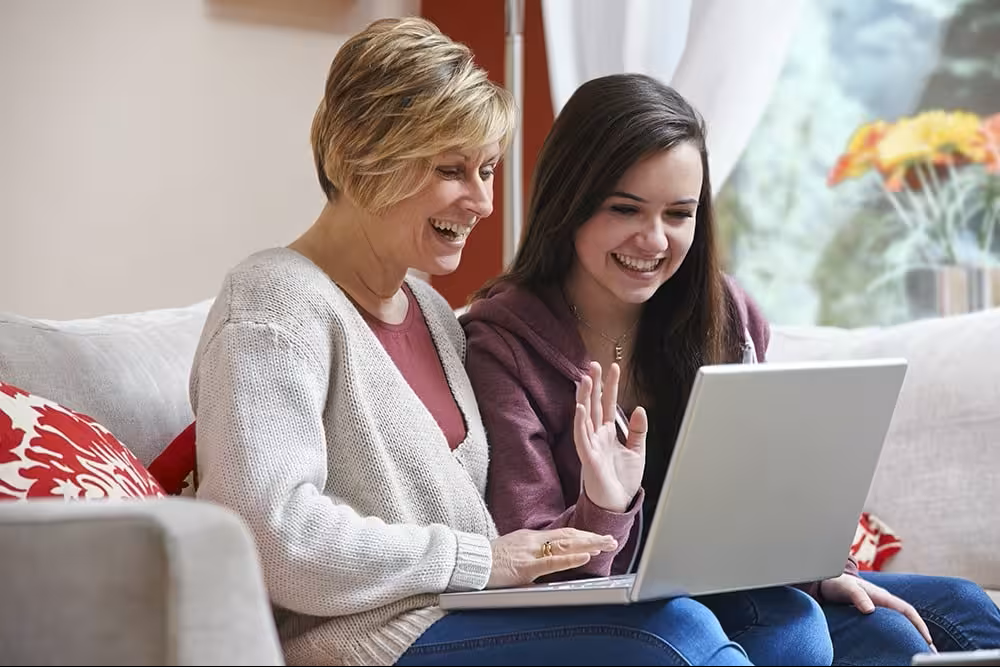 A cozy living room scene featuring two women sitting on a plush, light gray couch adorned with vibrant red-patterned cushions. The older woman, dressed in a soft cream cardigan, and the younger woman, in a burgundy hoodie, share a moment of joy as they engage with a silver laptop, sunlight illuminating the inviting space.