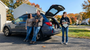 A gray crossover SUV with its rear hatch open sits on a suburban driveway, showing bags and packed items in the cargo area. Three adults stand beside it in casual fall clothing, with a gray house, green lawn, and autumn trees in the background under bright daylight.