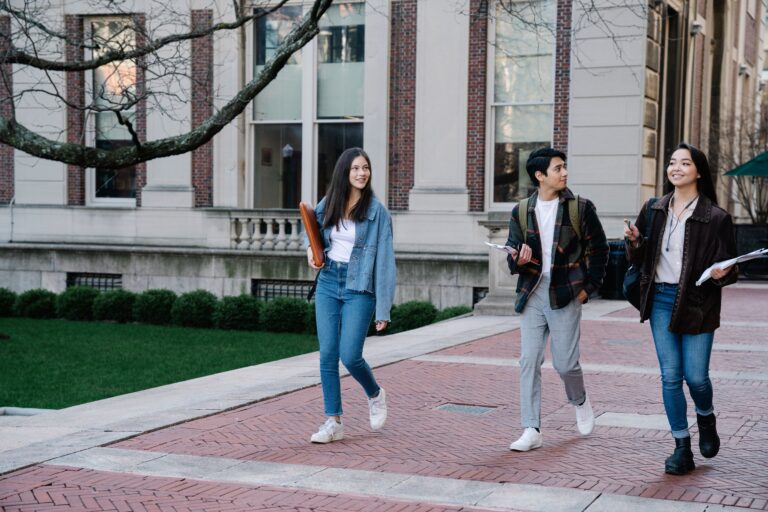 Three students walk together along a brick campus path beside a manicured lawn and a stone-and-brick building. They wear casual layers and jeans, with daylight softening the pale facade and bare tree branches framing the scene.