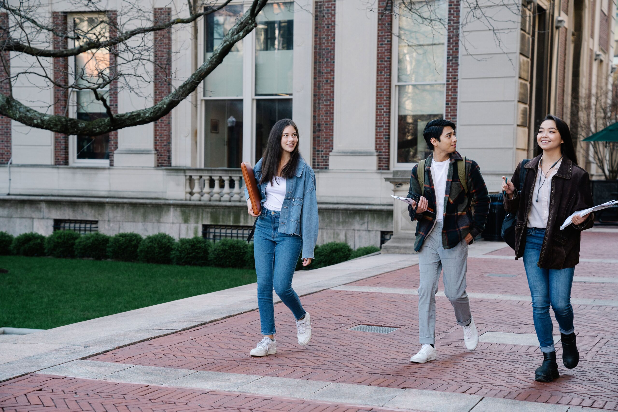 Three students walk together along a brick campus path beside a manicured lawn and a stone-and-brick building. They wear casual layers and jeans, with daylight softening the pale facade and bare tree branches framing the scene.