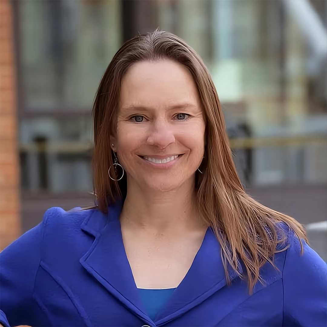 Headshot of a woman with shoulder-length brown hair wearing a royal blue blazer. She is smiling and leaning on a metal railing.