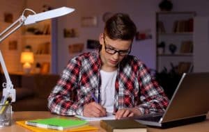 A teenage boy wearing glasses and a plaid shirt is sitting at a desk, writing in a notebook. A laptop, books, and notebooks are on the desk. The background shows shelves with books and a lamp.