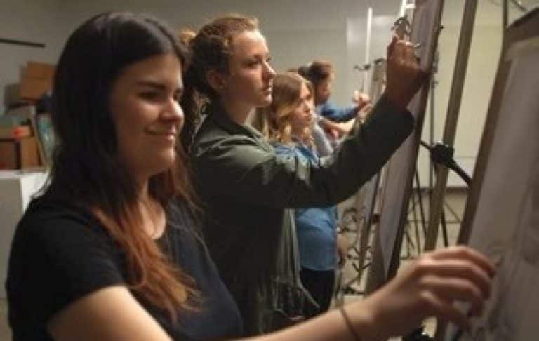 Several young women stand at easels, focused on drawing or sketching in an art class, with sketchpads and pencils in their hands. The background shows art supplies and a classroom setting.