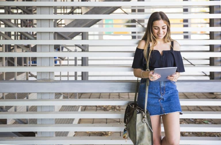 A young woman stands outside against a modern metal slat wall, smiling while looking at a tablet. She wears a black off-shoulder top, denim skirt, and carries a large bag over one shoulder.