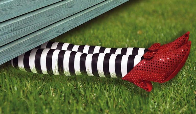 A pair of legs in black-and-white striped stockings and red sequined shoes, like a scene after SAT2 subject tests, stick out from under a light blue wooden structure on green grass.