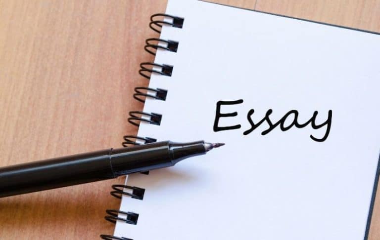 A black marker pen rests on an open spiral notebook with the word Essay written on a blank white page, placed on a wooden surface.