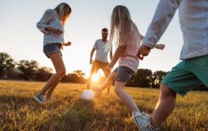 A family of four plays soccer together on a grassy field at sunset, smiling and enjoying the outdoors. The sun shines in the background, casting a warm glow over the scene.