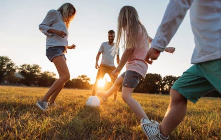 A family of four plays soccer together on a grassy field at sunset, smiling and enjoying the outdoors. The sun shines in the background, casting a warm glow over the scene.