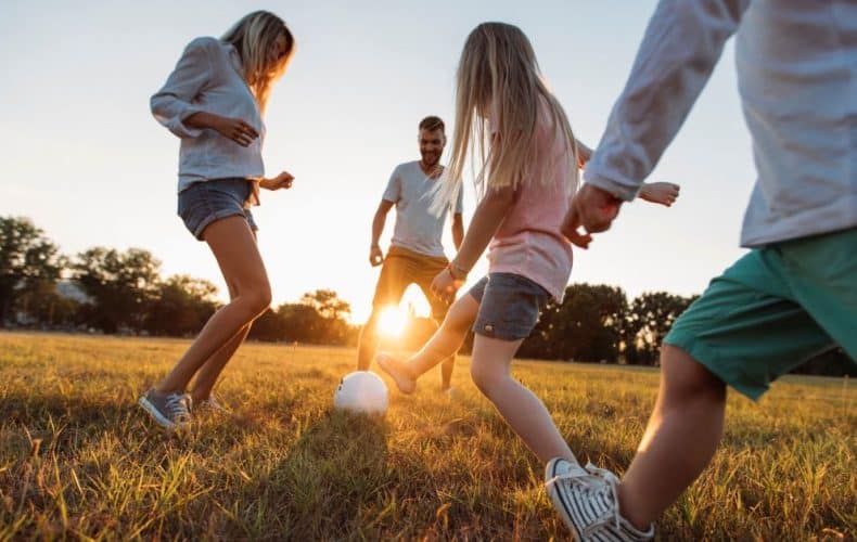 A family of four plays soccer together on a grassy field at sunset, smiling and enjoying the outdoors. The sun shines in the background, casting a warm glow over the scene.