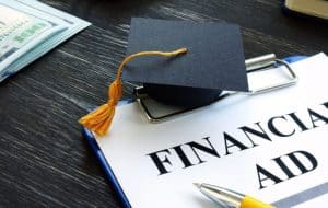 A small graduation cap sits on a clipboard with a paper labeled Financial Aid. A pen and a stack of cash are visible nearby on a dark wooden surface.