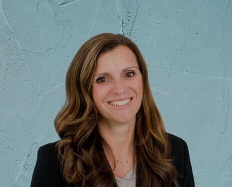 Headshot of a woman with shoulder-length brown hair, wearing a black blazer against a light blue textured background.