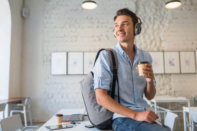 A young man wearing headphones and a backpack, possibly studying about American universities from Chile, sits on a table in a bright cafe, smiling and holding a takeaway coffee cup. A phone, notebook, and coffee cup are on the table beside him.