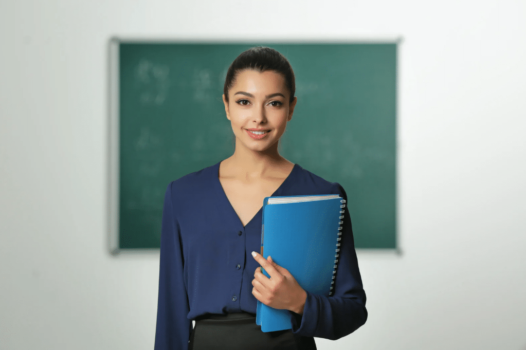 A woman stands in front of a chalkboard holding a blue notebook, smiling, dressed in a navy blouse and black skirt, suggesting a classroom or educational setting.