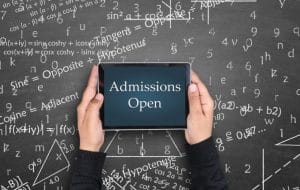 A person holds a tablet displaying Admissions Open in front of a chalkboard covered with math equations, formulas, and symbols.
