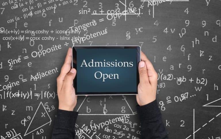 A person holds a tablet displaying Admissions Open in front of a chalkboard covered with math equations, formulas, and symbols.