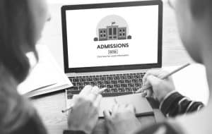 Two people sit at a table, using a laptop. The screen displays an admissions webpage with an illustration of a school building and a button labeled Enter. One person is holding a pencil and notebook.