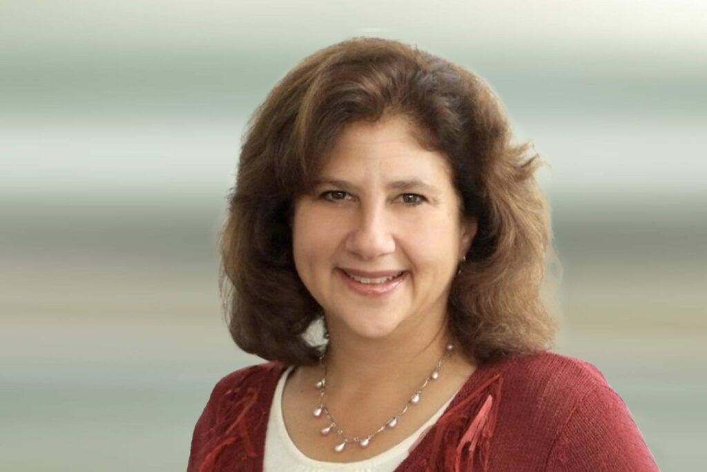 A woman with medium-length brown hair, wearing a red sweater over a white top and a beaded necklace, smiles at the camera against a blurred, light-colored background.