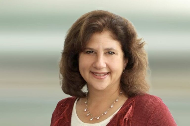 A woman with medium-length brown hair, wearing a red sweater over a white top and a beaded necklace, smiles at the camera against a blurred, light-colored background.
