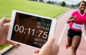 A person holds a tablet showing a stopwatch at 11.75 seconds on a running track, while a smiling runner in a pink shirt and race bib runs toward the finish line in the background.