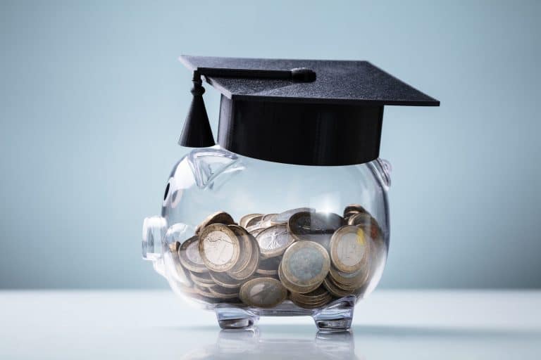 A clear piggy bank filled with euro coins wears a black graduation cap, symbolizing saving money for expensive private college education or student expenses, set against a plain light blue background.