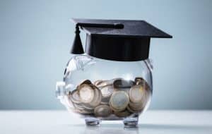 A clear piggy bank filled with coins is topped with a black graduation cap, symbolizing saving money for education or student financial planning.